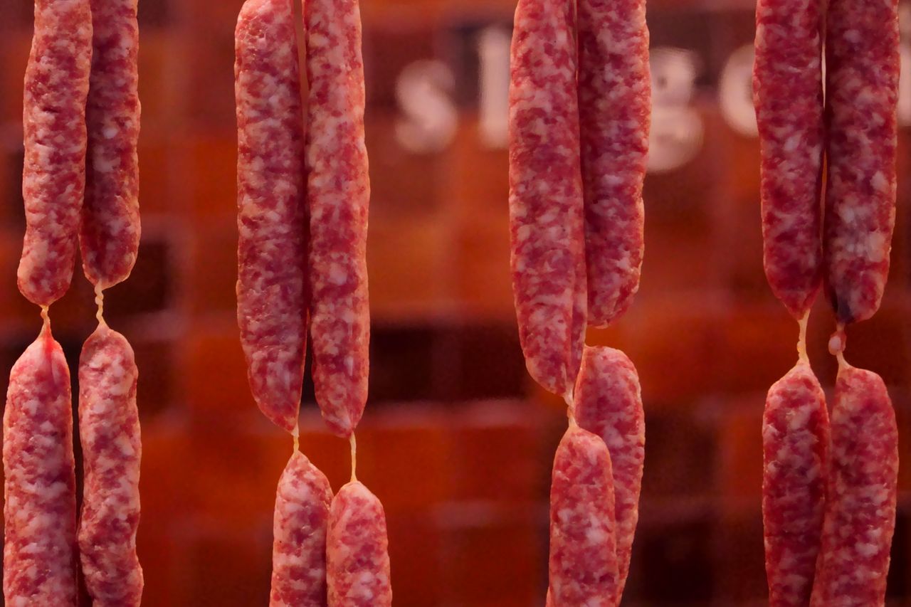 hero-img-02 Close-up of hanging cured sausages in a traditional butcher shop setting.