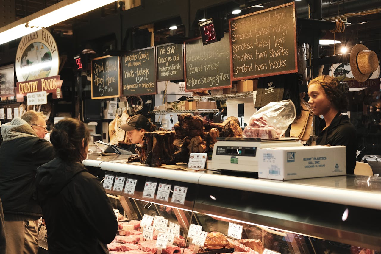about-01 A bustling meat shop showcasing various meats with employees serving customers over a counter.